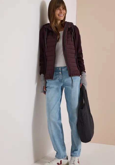 Lächelnde Frau trägt Jeans, graues Shirt, bordeauxfarbene Steppjacke und Tasche.