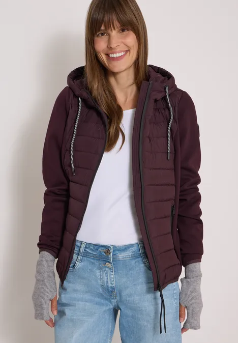 Eine lächelnde Frau trägt eine weinrote Steppjacke, weißes Shirt, Jeans und graue Handstulpen.