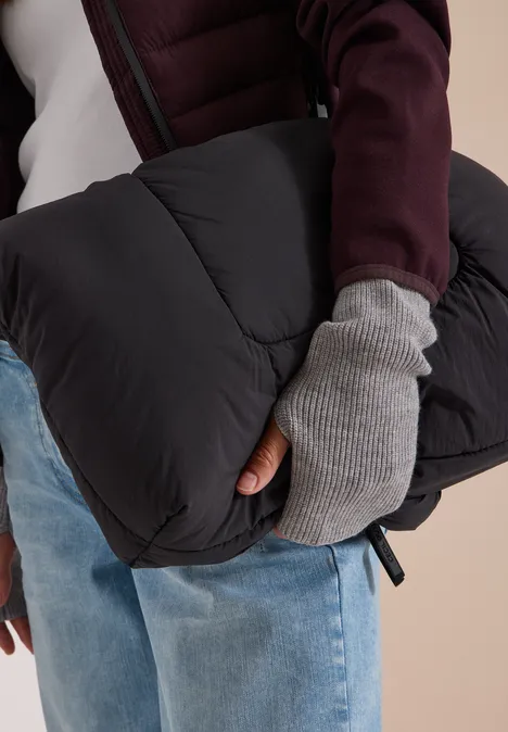 Person in Jeans und gestepptem Mantel mit grauen Fingerhandschuhen hält schwarze Pufferjacke.