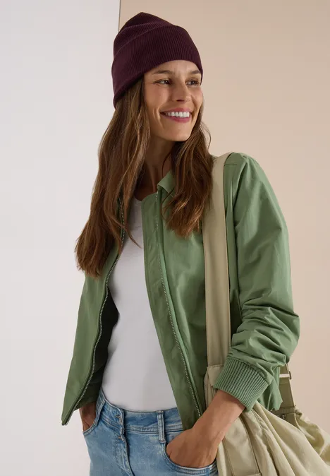 Eine lächelnde Frau mit grüner Jacke, weißem Top, Jeans, Beanie und Umhängetasche vor hellem Hintergrund.