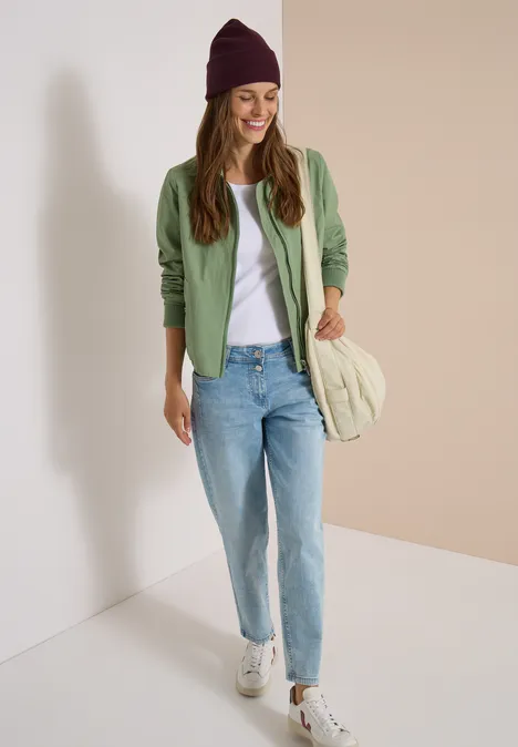 Junge Frau mit grüner Strickjacke, weißem T-Shirt, Jeans, Beanie und Sneakers lächelt in hellem Raum.