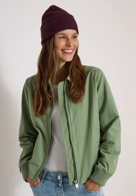 Lächelnde Frau in grüner Jacke, grauem Shirt und lila Mütze vor neutralem Hintergrund.