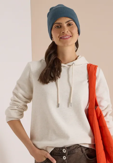 Frau in hellem Hoodie, blauer Mütze und roter Jacke über der Schulter, die lächelt.