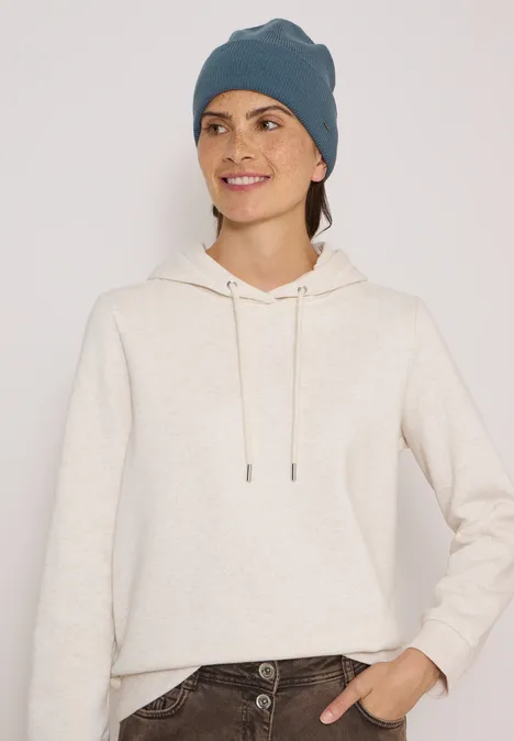 Frau mit beigem Hoodie und graublauer Strickmütze lächelt vor neutralem Hintergrund.