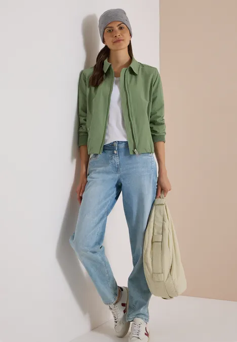 Frau in grüner Jacke, weißem Shirt, blauen Jeans und grauer Mütze hält eine beige Tasche.