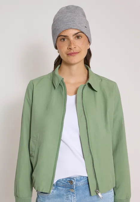 Junge Frau mit grauer Mütze, weißem T-Shirt und grüner Jacke vor schlichtem Hintergrund.