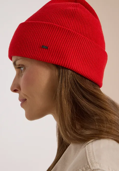 Seitenprofil einer Frau mit roter Strickmütze und heller Jacke vor hellem Hintergrund.