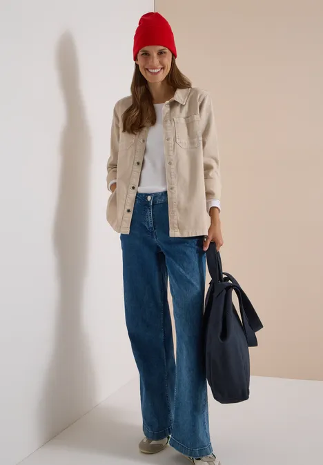 Frau mit roter Mütze, beiger Jacke, weißem Pullover und weiter Jeanshose trägt blaue Tasche.