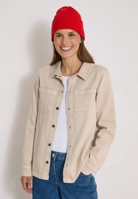 Junge Frau mit roter Mütze, beiger Jacke und weißem Shirt lächelt vor hellem Hintergrund.