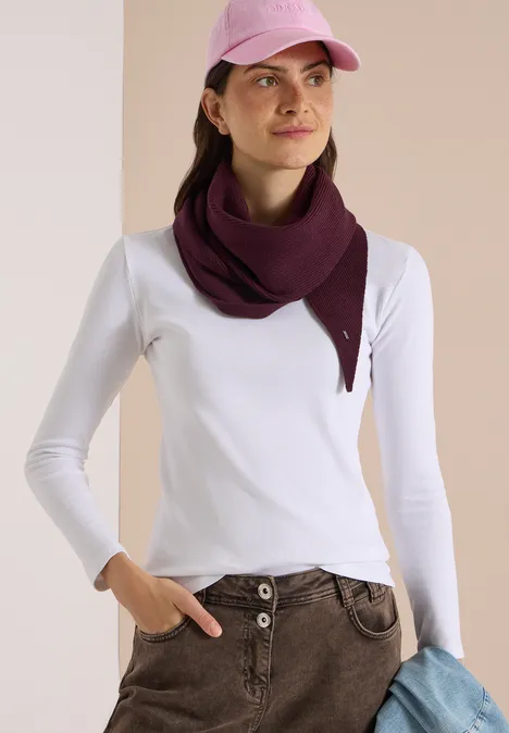 Frau mit weißem Langarmshirt, beiger Hose, violettem Dreiecksschal und rosa Cap, hält einen blauen Pullover