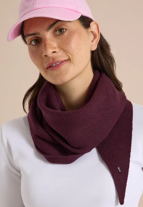 Frau mit rosa Baseballcap, violettem Schal und weißem Shirt vor neutralem Hintergrund.