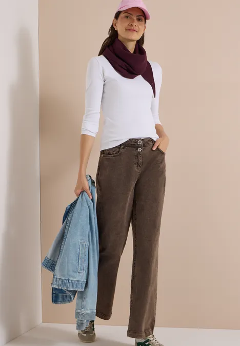 Frau trägt weiße Bluse, braune Hose, rosa Cap, hält Jeansjacke in der Hand, steht vor neutralem Hintergrund.