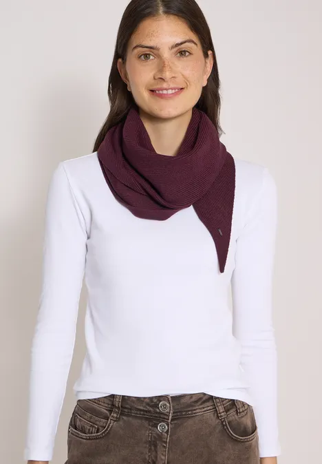 Frau lächelt, trägt weißen Langarmshirt und violetten Schal vor neutralem Hintergrund.