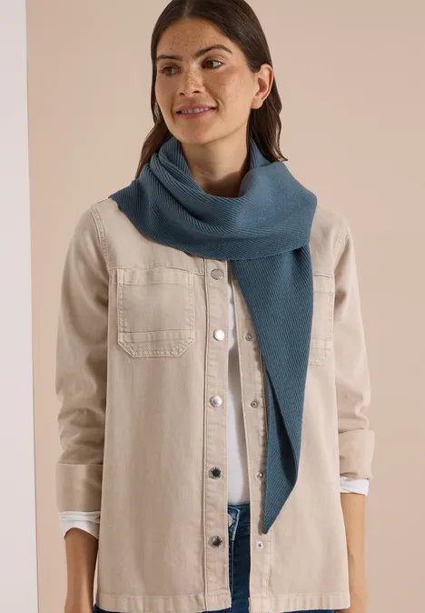 Frau mit heller Jacke und blauem Schal lächelt vor neutralem Hintergrund.