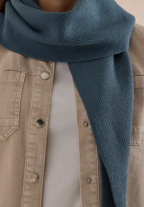 Blaue Strickschal um den Hals, dazu hellbeige Jacke und weißes Shirt.