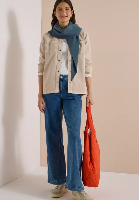 Frau mit heller Jacke, weißem Top, blauer weiten Jeans, blauem Schal und orangefarbener Stofftasche vor neutralem Hintergrund.