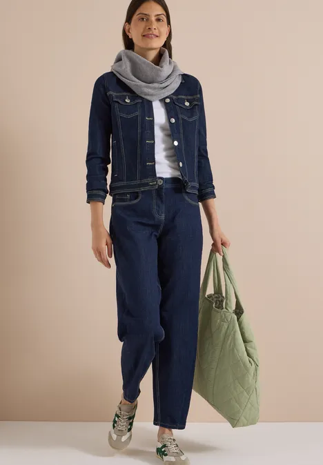 Frau in dunkelblauer Jeansjacke und Hose, grauem Schal, weißen Sneakern und großem grünen Shopper.