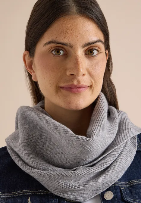 Frau mit braunen Haaren und grauem Strickschal, Denimjacke vor neutralem Hintergrund