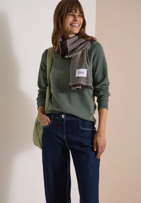 Vrouw in groene trui en sjaal, met jeans en tas, glimlachend tegen een neutrale achtergrond.