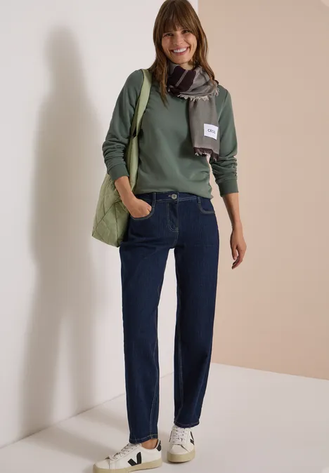 Een lachende vrouw in een groene trui, spijkerbroek, sjaal, tas en witte sneakers staat voor een neutrale achtergrond.