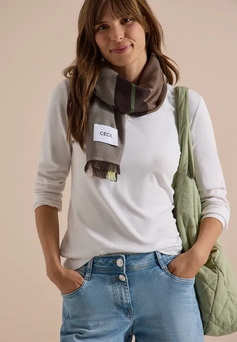 Jonge vrouw lacht in wit shirt, spijkerbroek, geruite sjaal en groene schoudertas.