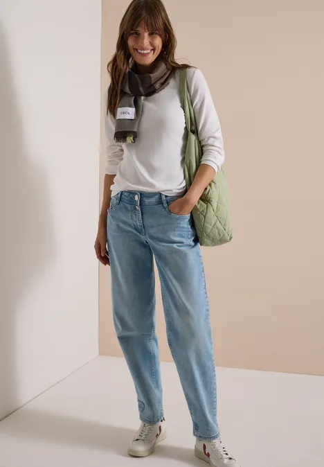 Jonge vrouw glimlacht in wit shirt, losse jeans, sjaal en groene tas voor een neutrale achtergrond.