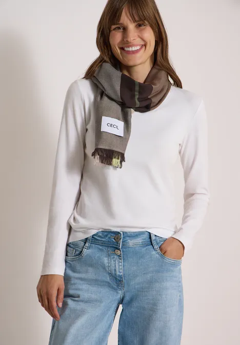 Een glimlachende vrouw draagt een wit shirt, blauwe jeans en een geruite sjaal.