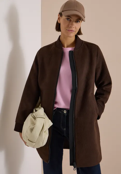 Frau in braunem Mantel, rosa Oberteil, dunkler Hose, beiger Tasche und Cap.
