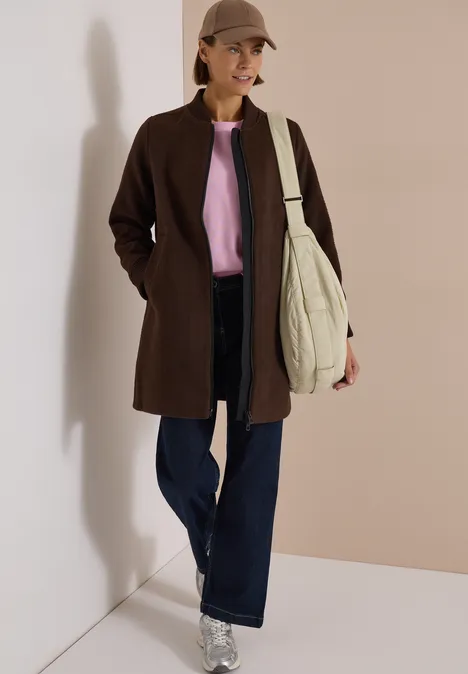 Frau mit braunem Mantel, rosa Pullover, weitem Jeans, Cap und großer Tasche vor neutralem Hintergrund.