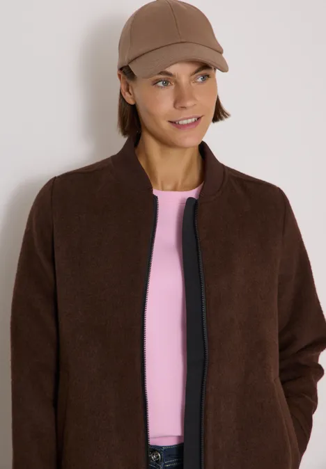 Frau trägt braune Jacke, rosa Shirt und Cap, lächelt.