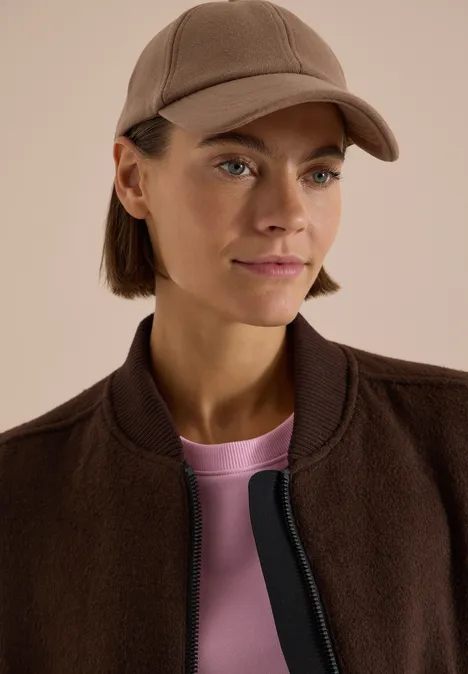 Frau mit brauner Baseballkappe, brauner Jacke und rosa T-Shirt, blickt seitlich.