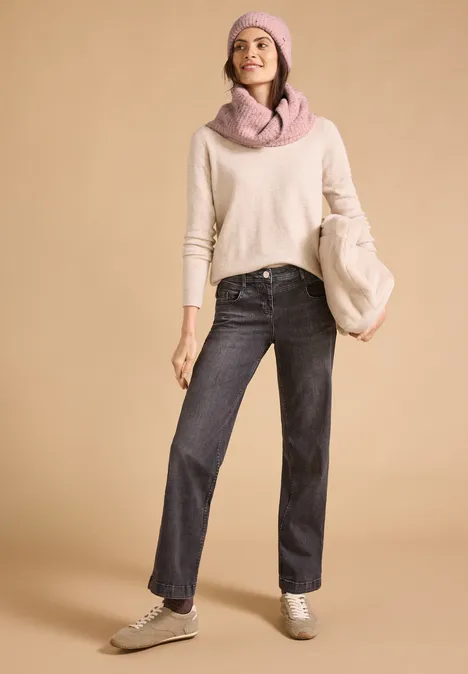 Junge Frau trägt hellen Pullover, schwarze Jeans, rosa Schal, Mütze und passende Tasche vor beigem Hintergrund.