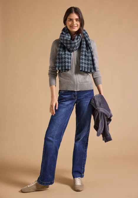 Kuscheliger Schal mit Fransen und Muster dark navy blue Frau in Jeans mit grauem Cardigan und blauem Schal hält Jacke und lächelt vor beigem Hintergrund.