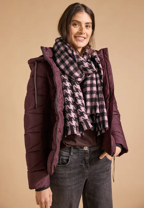 Kuscheliger Schal mit Fransen und Muster berry red Frau in lila Steppjacke und kariertem Schal, lächelt vor beige Hintergrund.