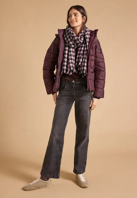 Kuscheliger Schal mit Fransen und Muster berry red Junge Frau in lila Steppjacke, kariertem Schal, dunklen Jeans und Sneakers posiert lässig.