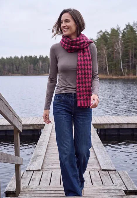 Zachte sjaal met franjes en patroon pink flash Vrouw loopt glimlachend op een steiger bij een meer, draagt grijs shirt, spijkerbroek en rood-zwart pied-de-poule sjaal.
