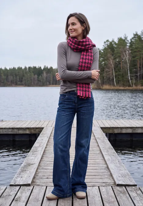 Zachte sjaal met franjes en patroon pink flash Een vrouw staat op een steiger bij een meer, in jeans, grijze trui en geruite sjaal