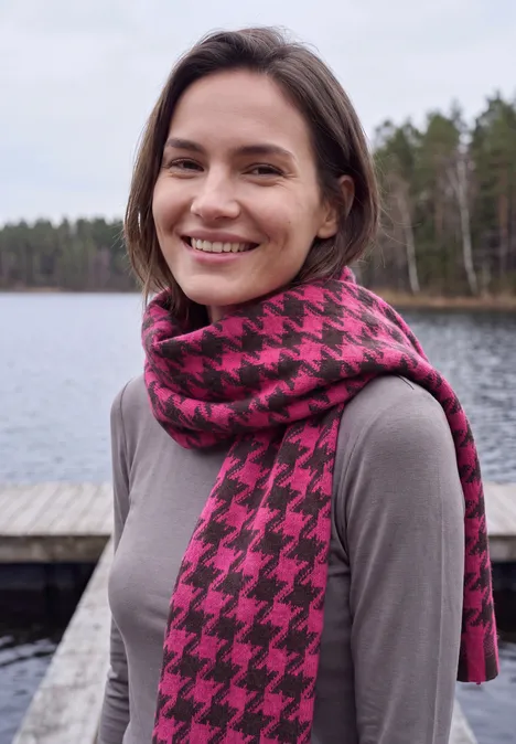 Zachte sjaal met franjes en patroon pink flash Vrouw lacht aan een meer, draagt grijs shirt en roze-zwart pied-de-poule sjaal, bos op de achtergrond.