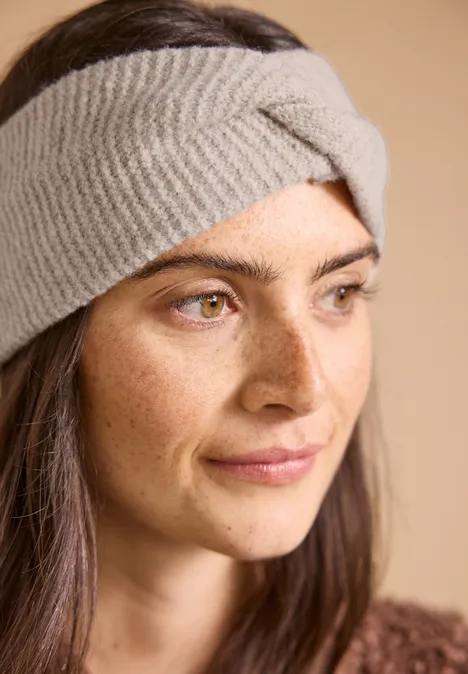 Eine Frau mit hellen Augen und braunen Haaren trägt ein graues gestricktes Stirnband und schaut sanft zur Seite.