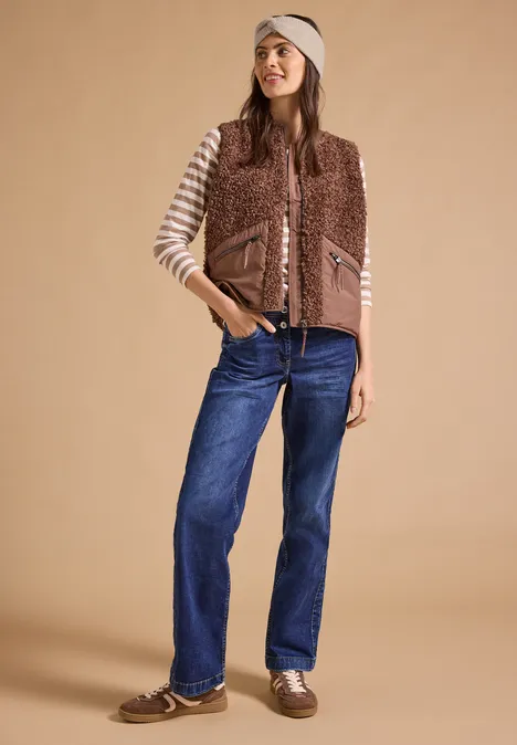 Frau mit gestreiftem Langarmshirt, brauner Fleeceweste, blauen Jeans und braunen Schuhen vor beigem Hintergrund.