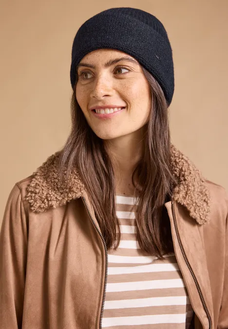 Frau mit schwarzer Strickmütze, brauner Jacke mit Teddy-Kragen und gestreiftem Shirt vor beigem Hintergrund.