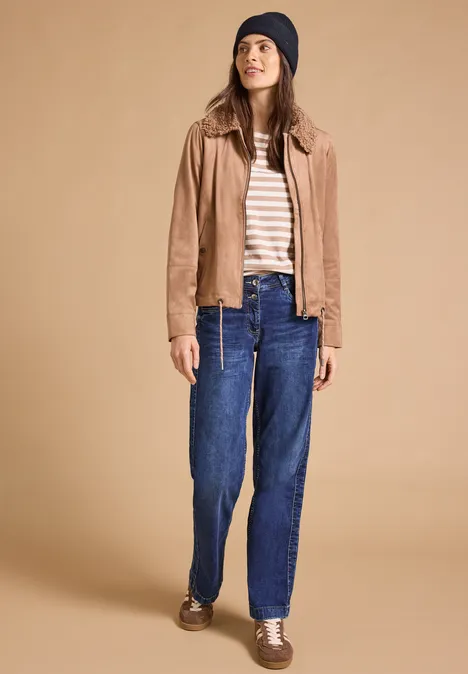 Frau mit Beanie, gestreiftem Shirt, beiger Jacke, blauen Jeans und braunen Schuhen vor schlichtem Hintergrund.