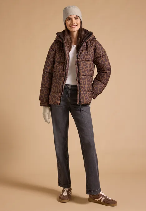 Frau in gemusterter brauner Steppjacke mit grauer Mütze und Jeans vor beigem Hintergrund.