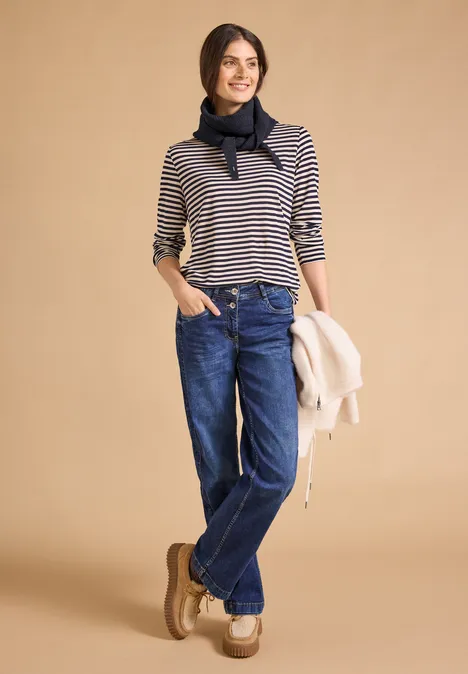 Eine Frau trägt eine gestreifte Bluse, Jeans, Halstuch, Halbschuhe und hält eine Jacke lässig in der Hand.