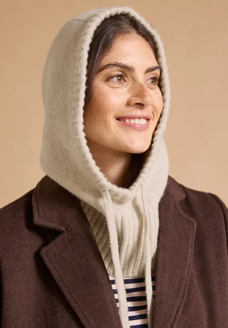 Balaclava mit Tunnelzug powder beige melange Frau mit cremefarbener Strickkapuze und braunem Mantel lächelt seitwärts.
