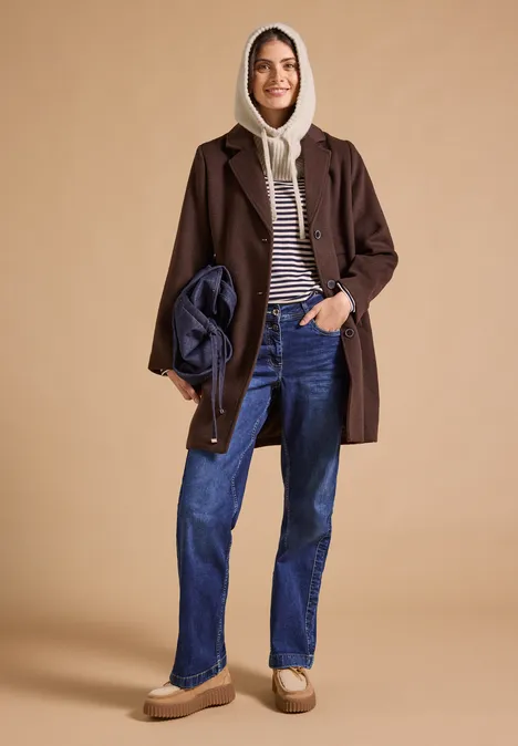 Balaclava mit Tunnelzug powder beige melange Frau lächelnd in braunem Mantel, Hoodie, Streifenshirt, blauen Jeans und beigen Schuhen, hält eine gesteppte Tasche.