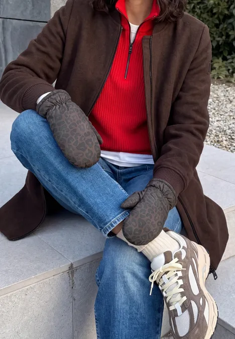 Gevoerde wanten met luipaardprint Black Person sitzt draußen auf einer Steinbank, trägt braunen Mantel, rotes Oberteil, graue Fäustlinge, Jeans und weiße Sneaker.