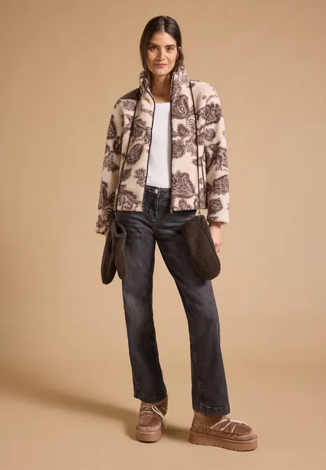 Fäustlinge im Teddy-Look chocolate sauce brown Frau in gemusterter Jacke, weißem Top, dunklen Jeans und braunen Fellschuhen vor beigem Hintergrund.