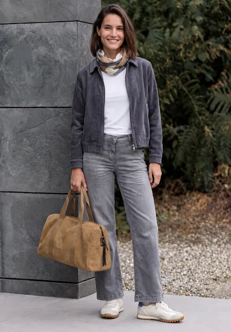 Frau in grauem Outfit mit Halstuch trägt eine braune Reisetasche und lächelt vor grauer Wand und Pflanzen.