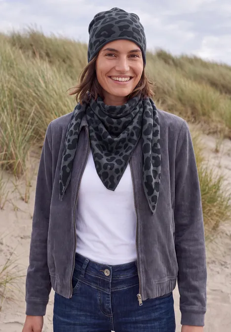 Personne debout sur une plage de dunes, porte t-shirt blanc, veste grise, écharpe et bonnet à motifs, sourit.
